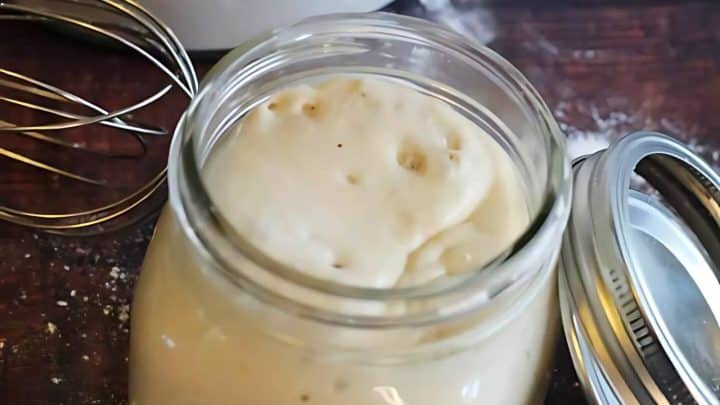 An open jar of creamy sourdough starter on a wooden table with a metal whisk nearby.