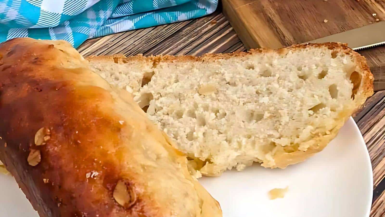Two slices of freshly baked bread on a white plate, with a knife and cutting board nearby. A blue checkered cloth is partially visible in the background.