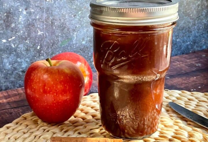 A mason jar with apple butter, apples and a knife next to it.