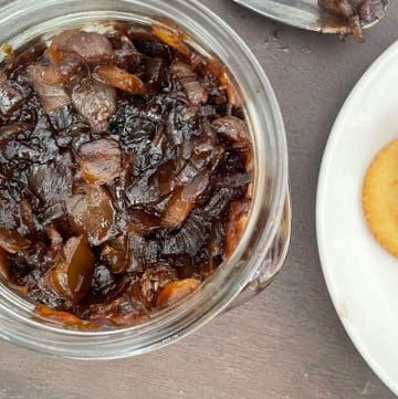 A jar of dark balsamic onion relish sits next to a plate with crackers generously topped with the same rich jam, all elegantly placed on a rustic wooden surface.