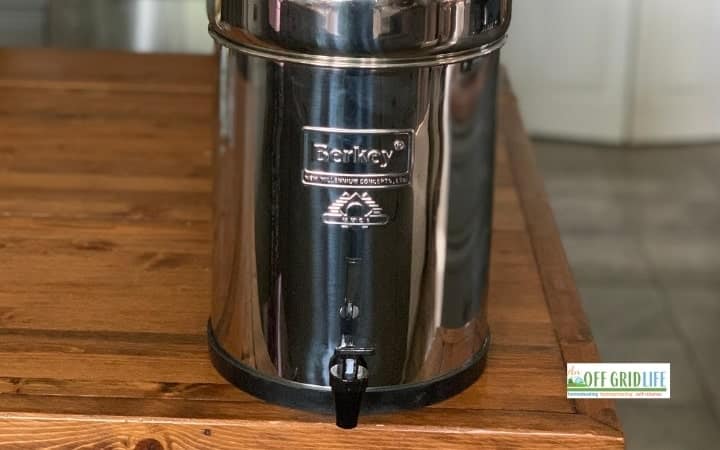 A stainless steel Berkey water filter dispenser, perfect for eco friendly gifts, sits on a wooden table with a faucet at the bottom and a logo on the front.