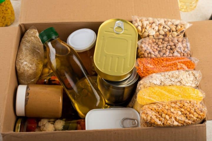 A cardboard box containing best foods for prepping such as bottles, cans, rice, lentils, chickpeas, oats, cornmeal, and jars.