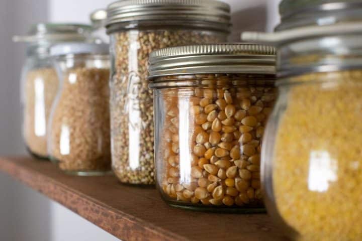 Glass jars filled with various grains, including corn kernels and seeds, are neatly arranged on a wooden shelf.