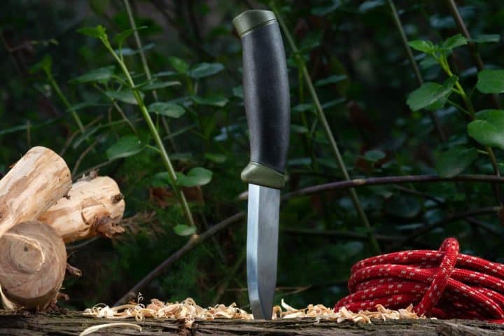 A survival knife stands upright on a wood log, surrounded by small wood shavings, chopped logs, and a coil of red rope against a leafy backdrop.