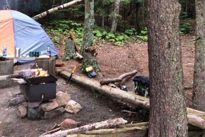 A campsite in a forest with a basic tent camping gear including tent, a log bench, a small campfire burning in a grill, and various camping supplies.