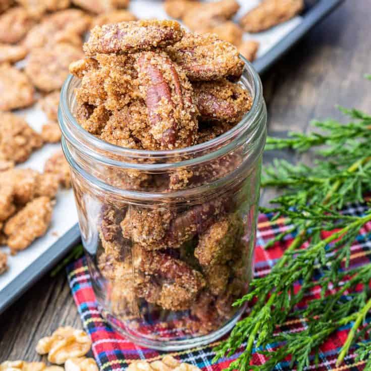 A glass jar filled with candied pecans sits on a tartan cloth, making perfect homemade food gifts. More candied pecans rest on a baking tray, surrounded by touches of greenery.