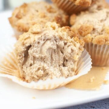 A halved sourdough discard muffin with crumbled topping graces a white plate, accompanied by three whole muffins in the background. The muffin reveals a delightfully crumbly interior.