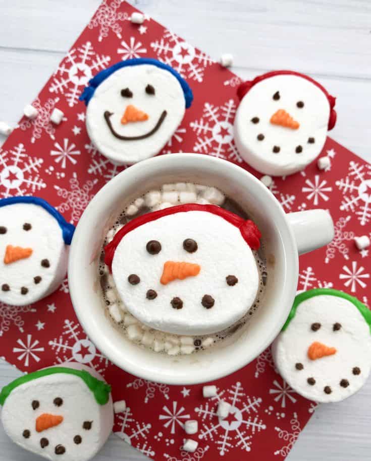 A mug of hot chocolate topped with a snowman-shaped marshmallow, surrounded by more snowman marshmallows on a red snowflake-patterned surface—perfect for homemade food gifts.