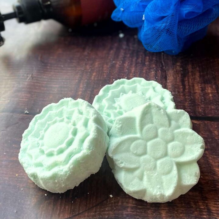 Three green aromatherapy shower steamers on a table next to a bottle of soap.