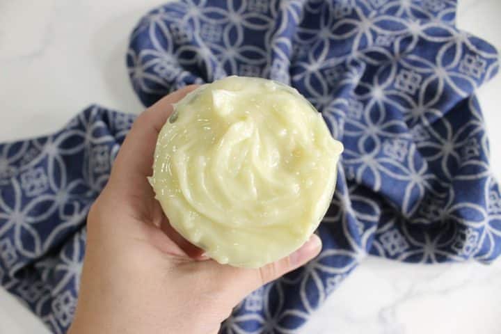 A hand cradles a jar filled with creamy, pale yellow DIY hair butter, set against a backdrop of blue patterned cloth.