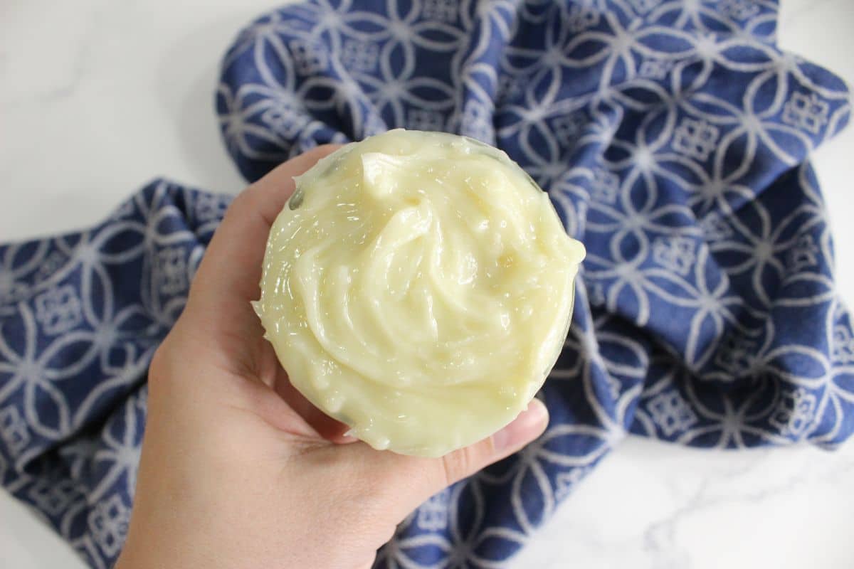 A hand cradles a jar filled with creamy, pale yellow DIY hair butter, set against a backdrop of blue patterned cloth.