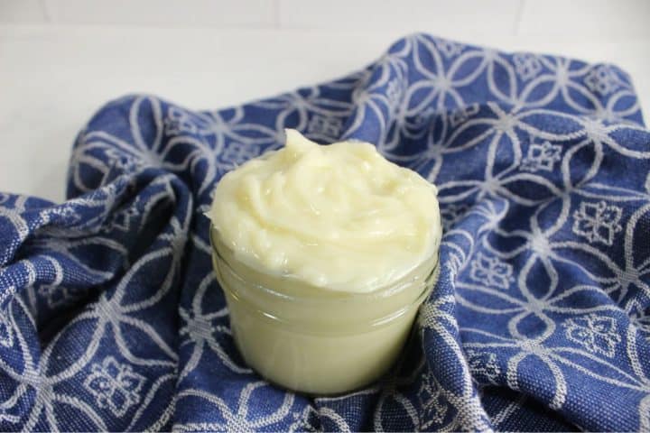 A small glass jar filled with DIY hair butter, is placed on a blue patterned cloth.