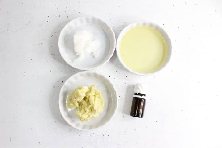 Small bowls with shea butter, coconut oil, and olive oil are arranged with a small dark bottle on a white surface.