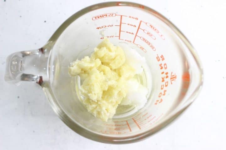 A glass measuring cup contains solid coconut oil and shea butter on a white surface.
