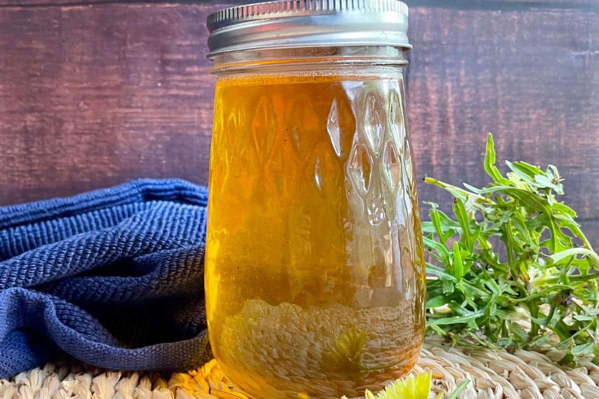 A jar of golden dandelion jelly with a metal lid on a woven mat next to fresh herbs and a blue cloth.