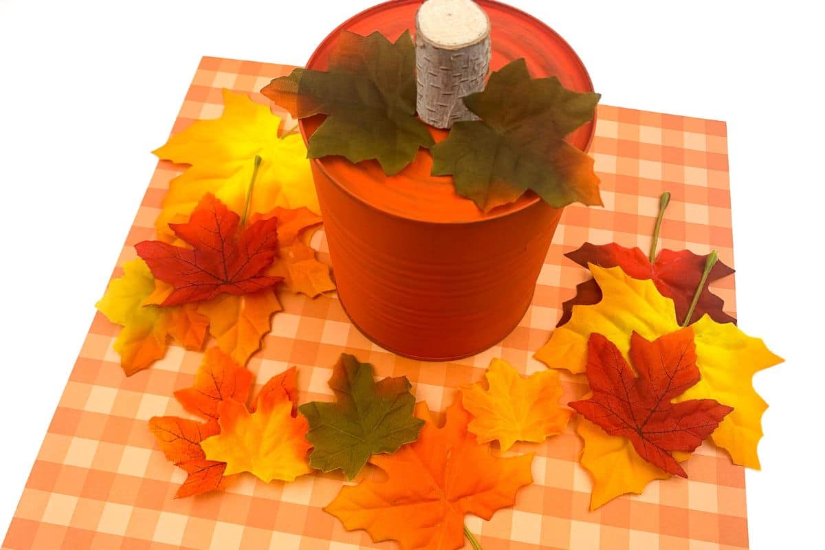 Orange can decorated to resemble a pumpkin, topped with a cork and surrounded by autumn leaves on a checkered surface.