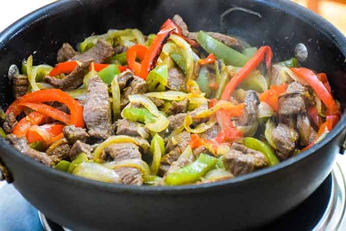 Sliced beef, red and green bell peppers, and onions cooking in a black skillet on a stovetop—a tasty dish perfect for those seeking venison dinner ideas.