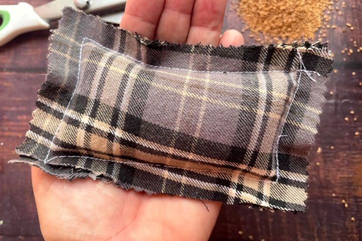 A small rectangular plaid sachet eco-friendly hand warmers filled with grains is held in a hand over a wooden surface. Scissors and scattered grains are visible in the background.