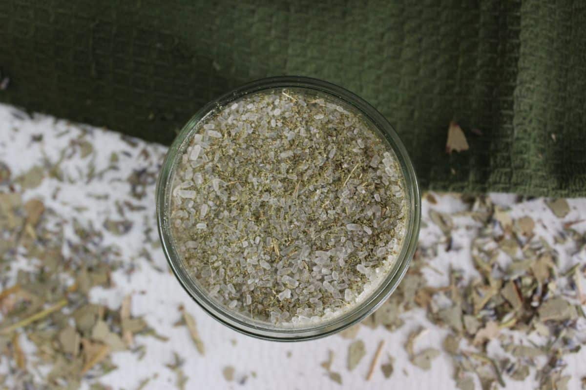 Glass jar filled with eucalyptus bath salts on a white surface with scattered herbs and a green cloth in the background.