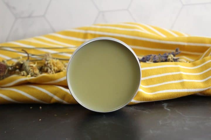 A tin of solid healing salve is placed on a black surface, in front of a yellow and white striped cloth, with dry herbs scattered around.