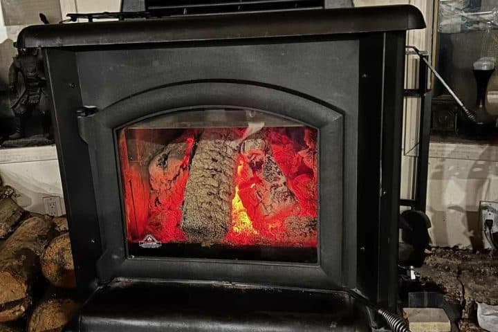 A black wood-burning stove with visible logs glowing red inside, placed indoors near a pile of firewood.