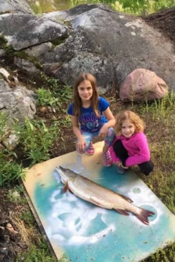 two young girls outdoors next to a wooden plank with one large fish and fishing gifts