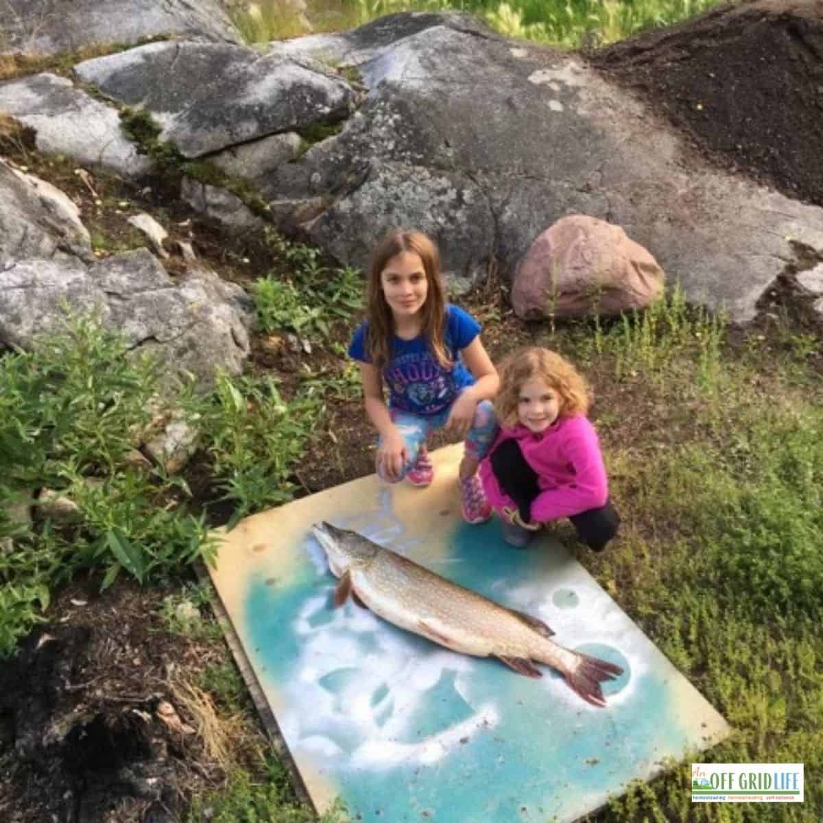 two young girls outdoors next to a wooden plank with one large fish and fishing gifts
