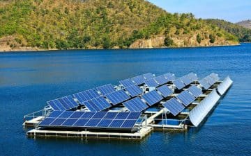 Floating solar panels on a calm body of water, surrounded by tree-covered hills under a clear blue sky.
