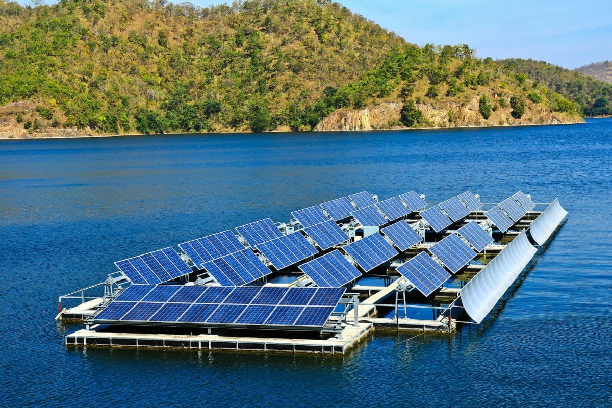 Floating solar panels on a calm body of water, surrounded by tree-covered hills under a clear blue sky.