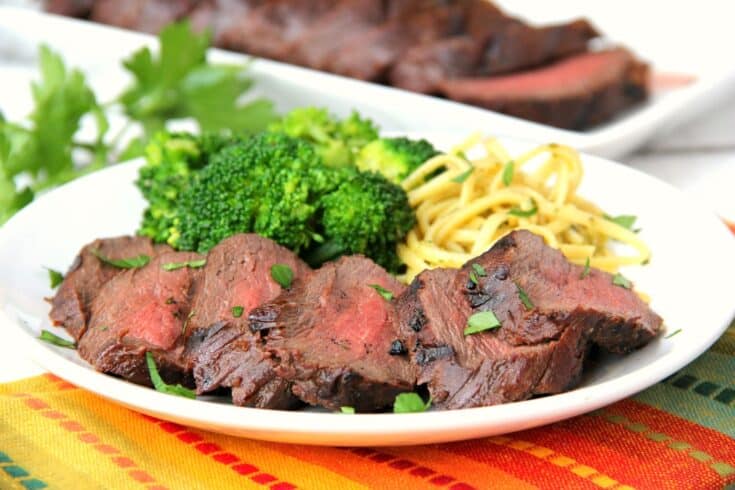 Sliced grilled steak, inspired by venison dinner ideas, served with steamed broccoli and a portion of pasta on a white plate, garnished with fresh parsley.