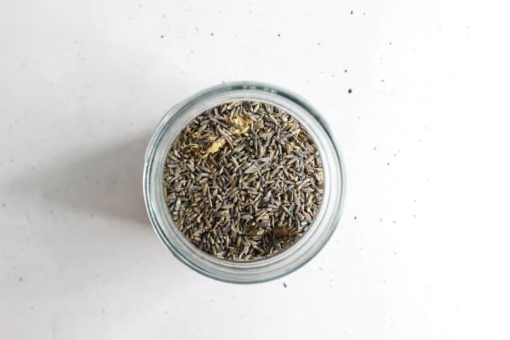 A glass jar filled with dried lavender buds viewed from above on a white surface.