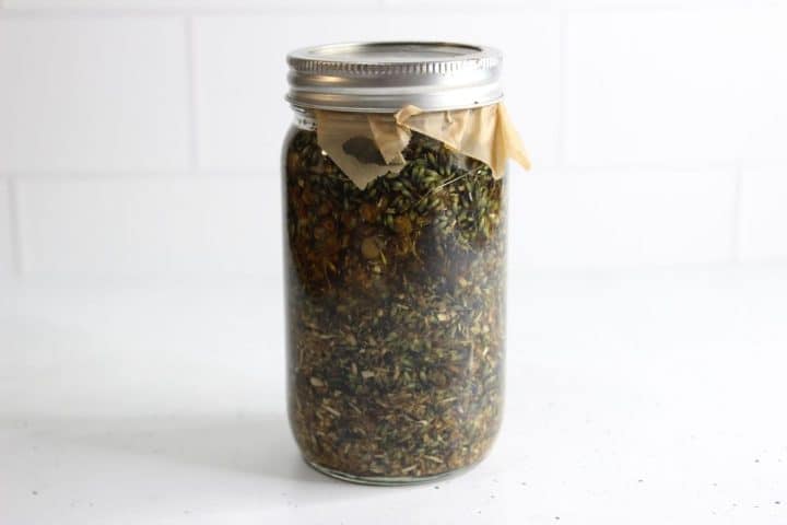 A glass jar filled with dried herbs and covered with a metal lid, placed on a white surface.