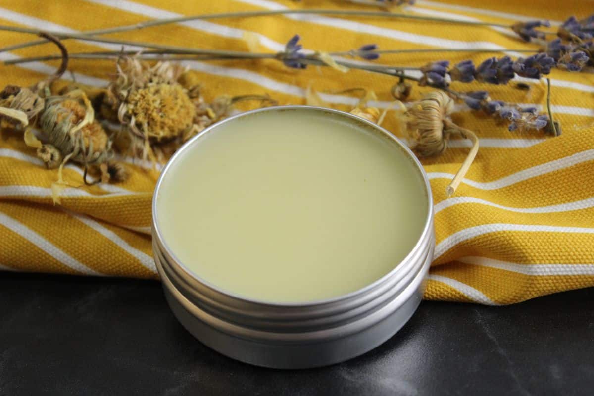 A tin of balm with a smooth surface sits on a yellow striped cloth, surrounded by dried lavender and other herbs.