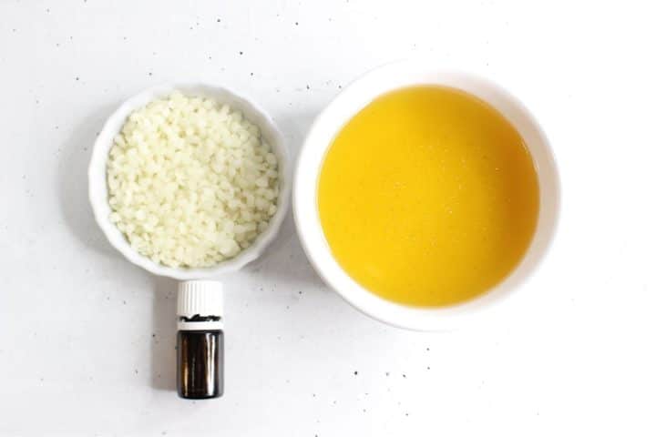 Small bottle of essential oil next to a white bowl of beeswax pellets and a bowl of melted yellow beeswax on a white surface.