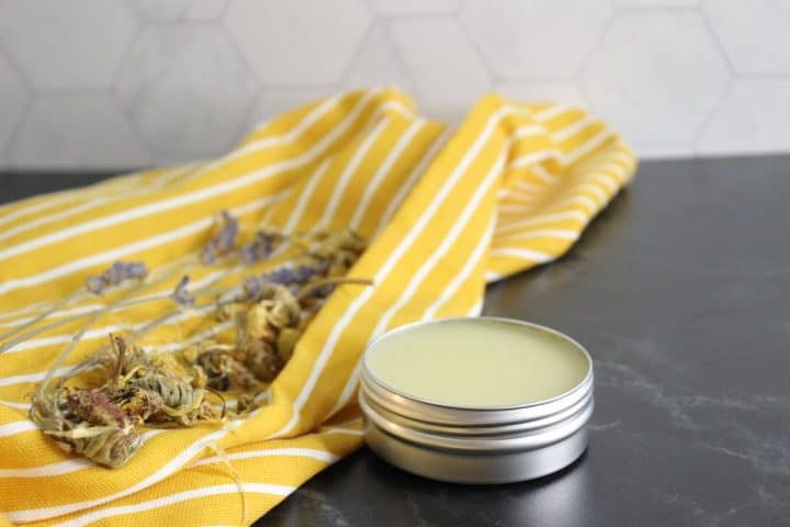 A tin of balm sits on a dark surface next to dried flowers on a yellow and white striped cloth.