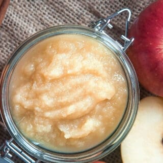 Homemade applesauce in a mason jar on a wooden tabletop with a sliced apple and cinnamon sticks