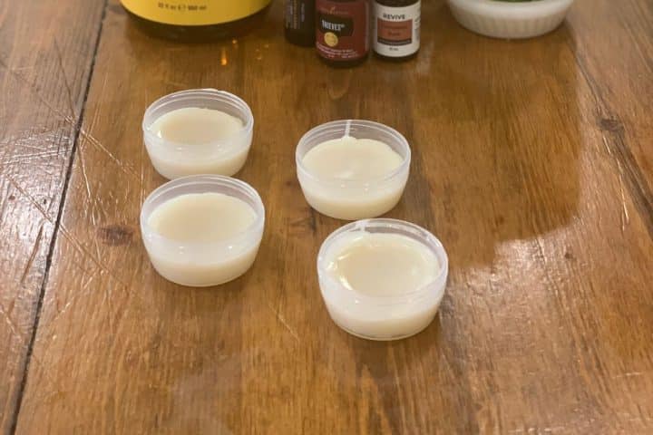 Four small plastic containers filled with orange spiced beeswax lip balm are placed on a wooden table, with several bottles of ingredients seen in the background.