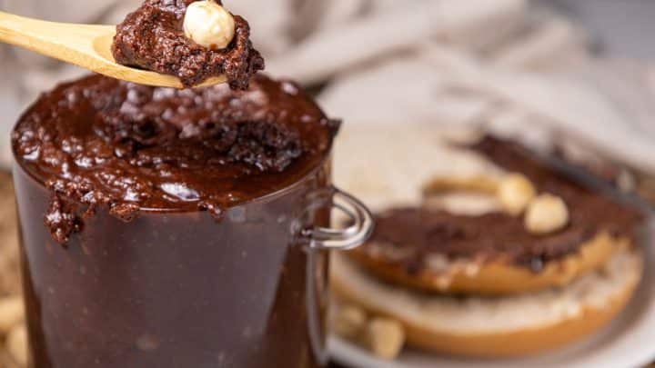 A spoonful of chocolate hazelnut spread being lifted from a glass jar, with a bagel covered in the spread in the background.