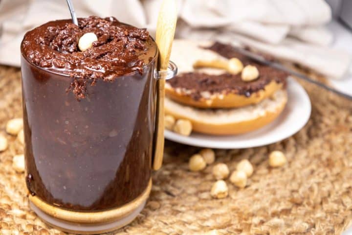 A glass jar filled with chocolate spread and a spoon is on a woven surface. A plate with bread and chocolate spread, surrounded by hazelnuts, is in the background.