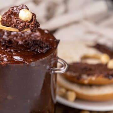 A spoon scoops chocolate hazelnut spread from a glass jar. A bagel spread with the same topping is on a plate in the background.