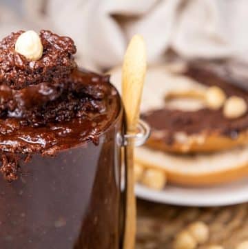 A spoonful of homemade Nutella with a hazelnut is held above a jar. In the background, a bagel is spread with the same rich chocolate and topped with crunchy hazelnuts.