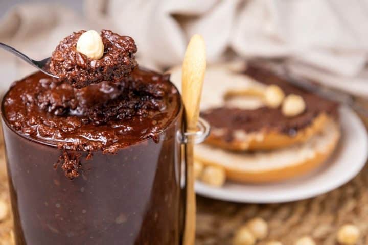 A spoonful of chocolate spread with a hazelnut over a glass jar, with bread topped with the same spread and hazelnuts in the background.