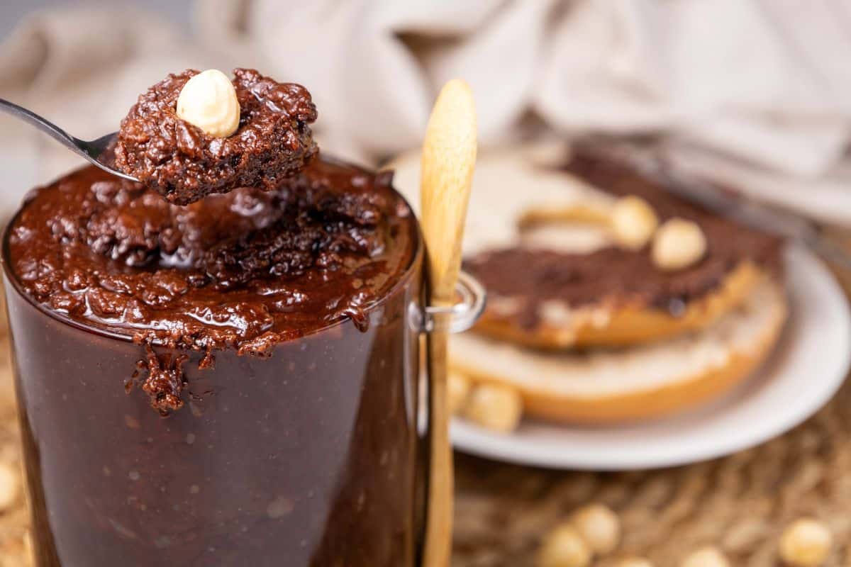 A spoonful of homemade Nutella with a hazelnut is held above a jar. In the background, a bagel is spread with the same rich chocolate and topped with crunchy hazelnuts.