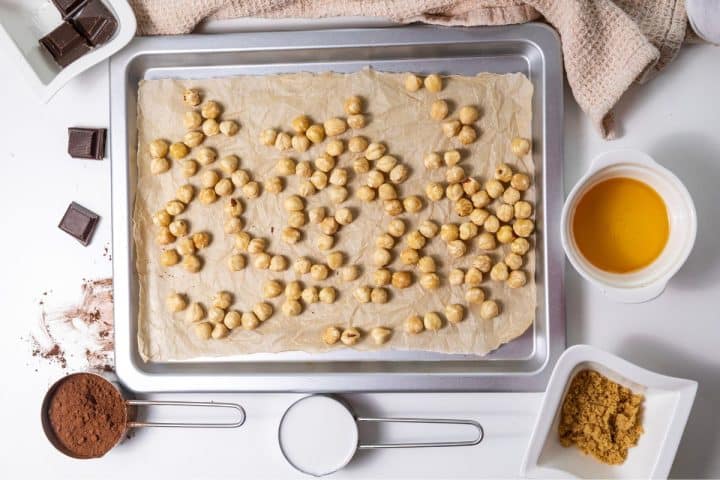 Hazelnuts spread on a baking sheet lined with parchment paper, surrounded by ingredients like chocolate, cocoa powder, sugar, and milk on a white surface.