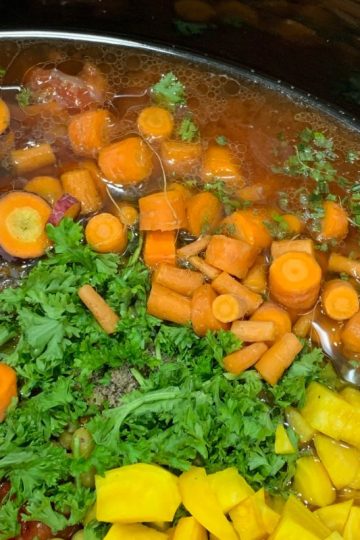 Chopped vegetables including carrots and yellow squash simmering in a slow cooker with herbs.