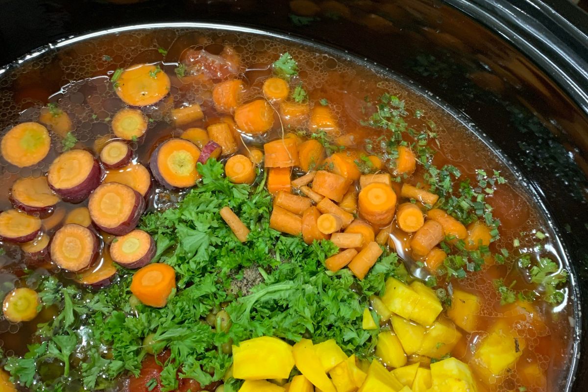 Chopped vegetables including carrots and yellow squash simmering in a slow cooker with herbs.