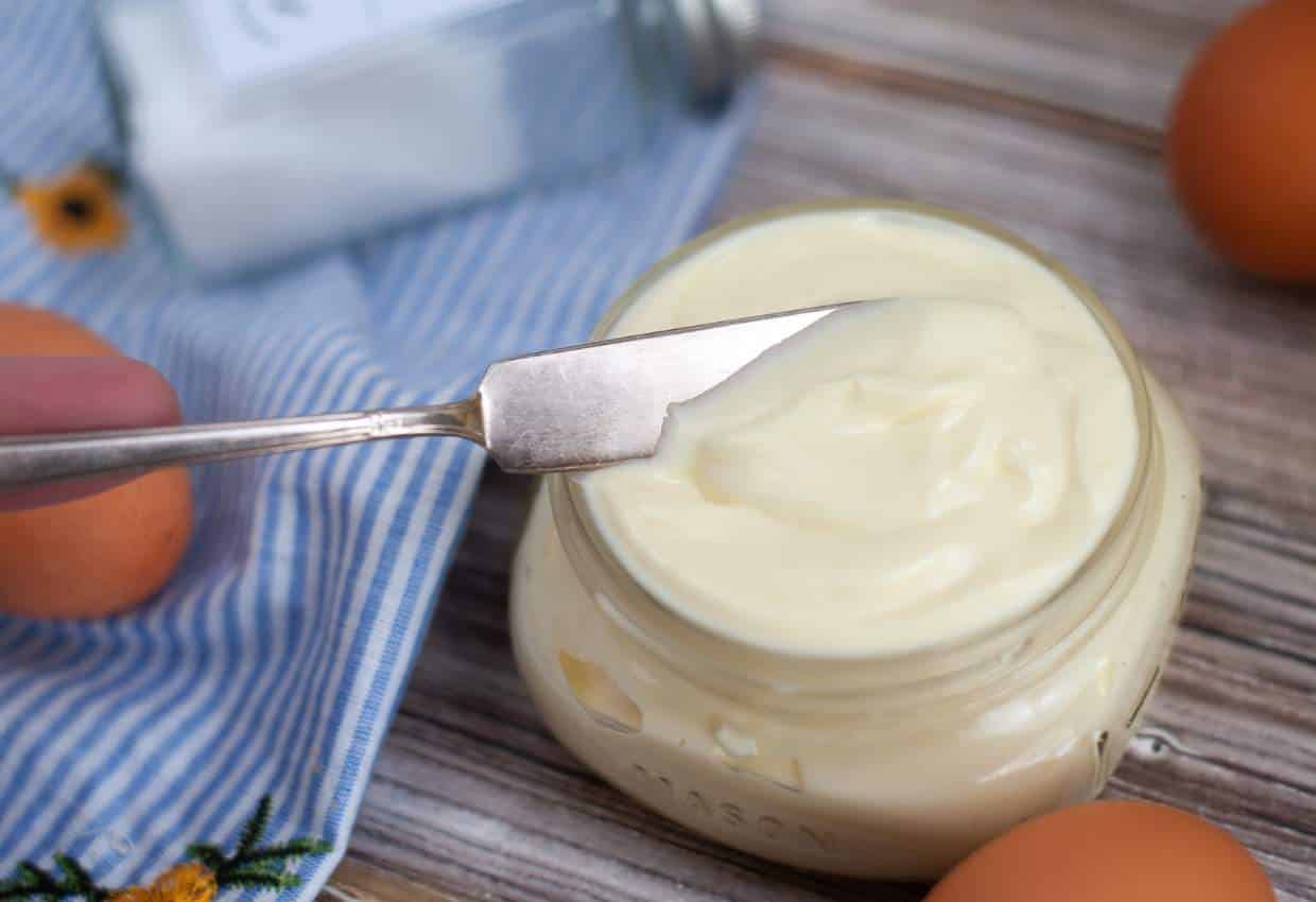 Homemade mayonnaise in a small mason jar with a knife, brown eggs, and cloth.