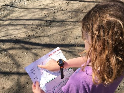 A young girl with wavy brown hair and a watch on her wrist looks at a map and points to a location while standing on a dirt path.