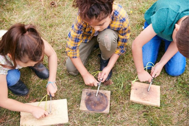 Three children kneel on the grass, learning to make fire using fire starter kits on wooden boards.