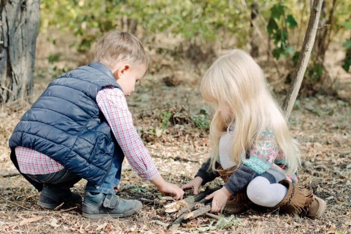 Two children, a boy and a girl, are crouched on the ground in a wooded area, arranging sticks and leaves. The boy wears a blue vest, and the girl has long blond hair and wears a knitted sweater.